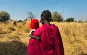 A woman carries a small child in a dry field, they are facing away from the camera. Wooden huts are visible in the background.