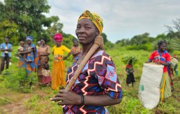 A woman stands in a field proudly carrying a wooden axe. Several other women stand behind her in the background.