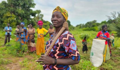 A woman stands in a field proudly carrying a wooden axe. Several other women stand behind her in the background.