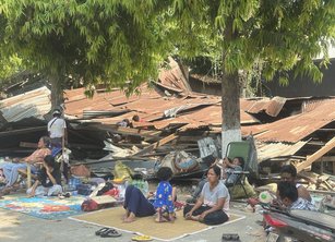 A family sit on a mat amidst buildings destroyed by the earthquake