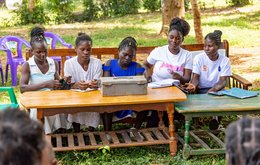 Five women sit together in a row on a bench at a desk, writing some notes