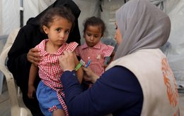 A female medic holds a measuring band around a young girl's arm. The young girl sits on a woman's lap, and another young girl watches on in the background.
