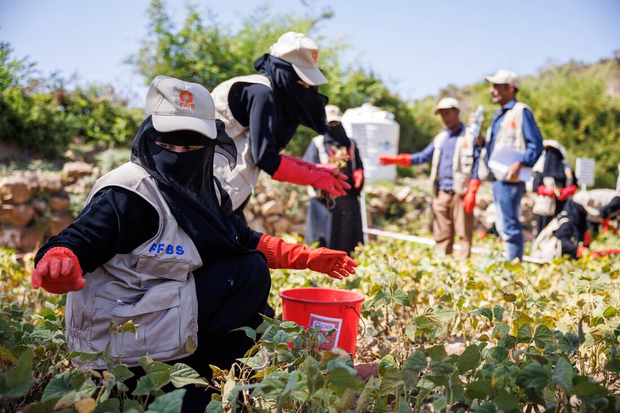 Two women working with crops in a field, both wearing CARE caps. Some other men and women are visible in the field in the background.