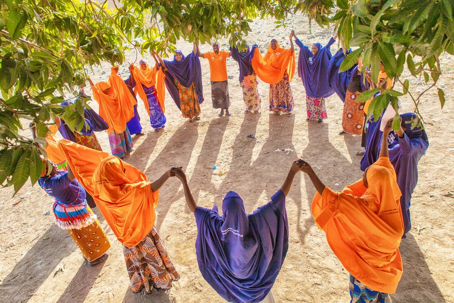 A group of about 20 women, all wearing either bright orange or vivid purple, stand together outside holding hands in a circle