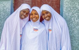 3 girls stand together smiling. They all wear white clothing with a badge that says 'March4Women' on it with the CARE logo