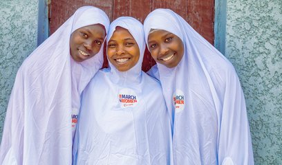 3 girls stand together smiling. They all wear white clothing with a badge that says 'March4Women' on it with the CARE logo