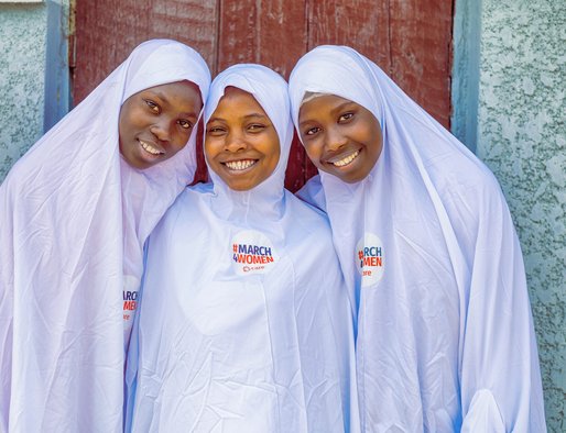3 girls stand together smiling. They all wear white clothing with a badge that says 'March4Women' on it with the CARE logo