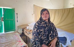 A woman looks to camera, sitting on a makeshift bed in a bare room with green shutters
