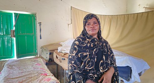 A woman looks to camera, sitting on a makeshift bed in a bare room with green shutters