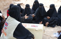 A group of about 10 women sit together on the floor at a meeting outside, led by a woman in a CARE vest