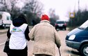 The back view of two people walking along a street arm in arm. One person is wearing a humanitarian vest, the other is older and walks with a stick.