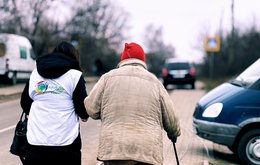 The back view of two people walking along a street arm in arm. One person is wearing a humanitarian vest, the other is older and walks with a stick.