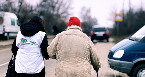 The back view of two people walking along a street arm in arm. One person is wearing a humanitarian vest, the other is older and walks with a stick.