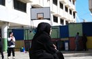 A woman sits in on a chair in the middle of what looks like a school playground. A child and a basketball hoop are in the background.