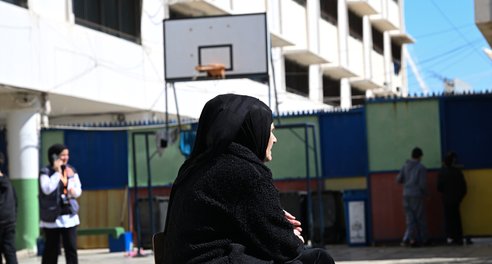 A woman sits in on a chair in the middle of what looks like a school playground. A child and a basketball hoop are in the background.