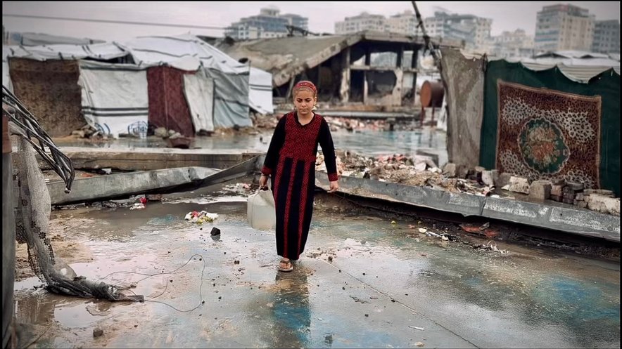A girl carries a jerrycan of water amidst some makeshift tents and rubble