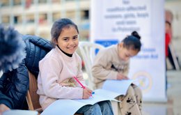 A young girl sits outdoors on a chair. She is smiling to camera while writing in a notebook with a pink pencil. A couple of other children are visible in the background, also writing in notebooks.