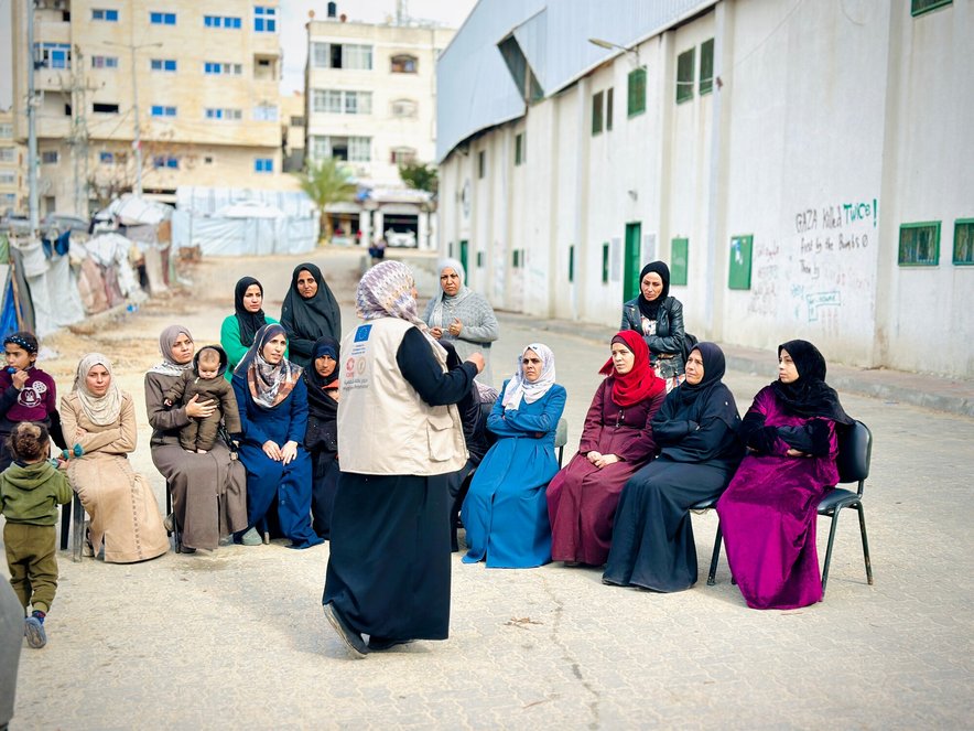 A woman talks to a group of about 12 women seated in a semi-circle outside a concrete block of flats