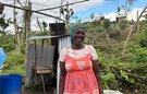 A woman smiles standing in front of a flimsy corrugated iron shelter and fallen down trees