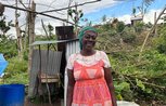 A woman smiles standing in front of a flimsy corrugated iron shelter and fallen down trees