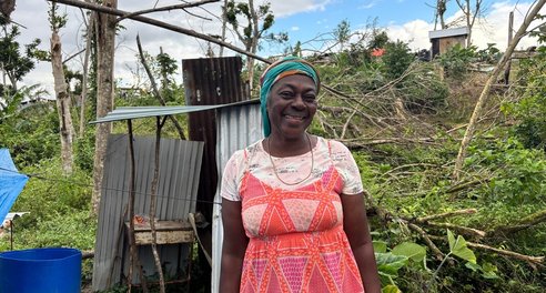 A woman smiles standing in front of a flimsy corrugated iron shelter and fallen down trees