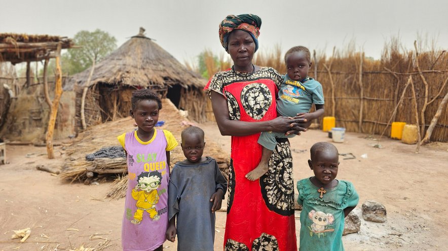 A woman stands in front of a straw hut with four young children