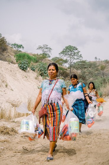 Three women walk along a dusty track carrying bundles of emergency hygiene supplies such as toothpaste, sanitary towels and toilet roll