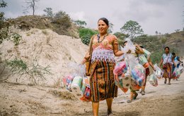 A group of women walk along a dusty track carrying large bundles of emergency hygiene supplies such as toothpaste, sanitary towels and toilet roll