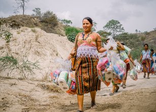 A group of women walk along a dusty track carrying large bundles of emergency hygiene supplies such as toothpaste, sanitary towels and toilet roll
