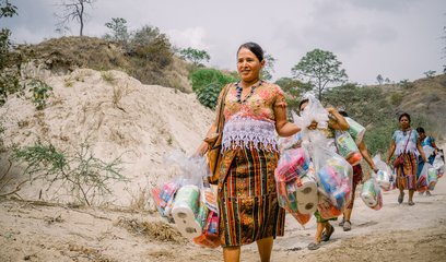 A group of women walk along a dusty track carrying large bundles of emergency hygiene supplies such as toothpaste, sanitary towels and toilet roll