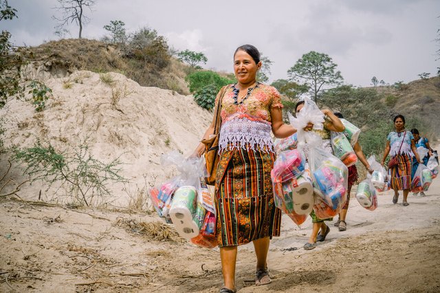 A group of women walk along a dusty track carrying large bundles of emergency hygiene supplies such as toothpaste, sanitary towels and toilet roll