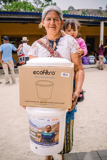 A woman with a small child on her back carries a large bucket and a cardboard box containing an eco-filter