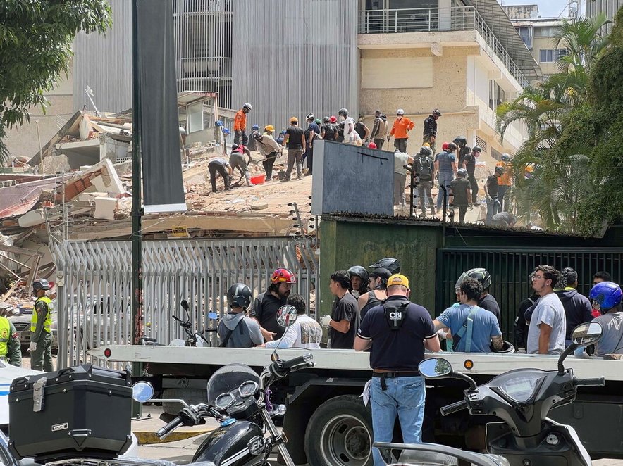 A group of rescue workers working through the rubble of a collapsed building. A group of onlookers are in the street in the foreground