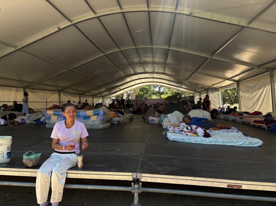A woman sits at the front of a large temporary shelter. Several people lying on mattresses are visible in the background.