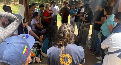 A woman in a CARE vest talks with a group of aid workers and earthquake survivors. Temporary shelters are visible in the background