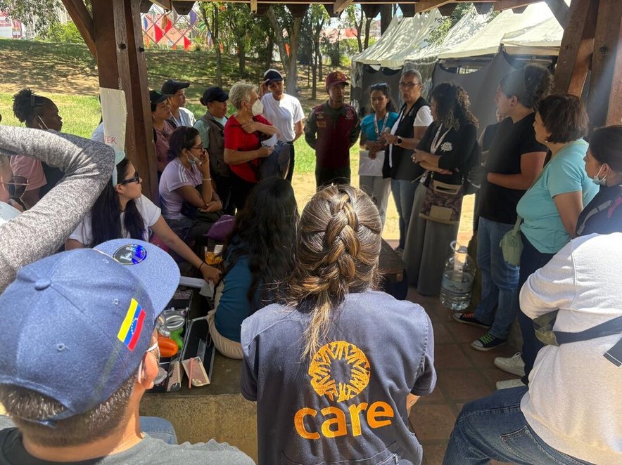 A woman in a CARE vest talks with a group of aid workers and earthquake survivors. Temporary shelters are visible in the background