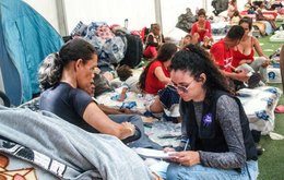 An aid worker talks with a woman in a temporary shelter. Many other earthquake survivors are visible in the background, sitting on mattresses on the floor.