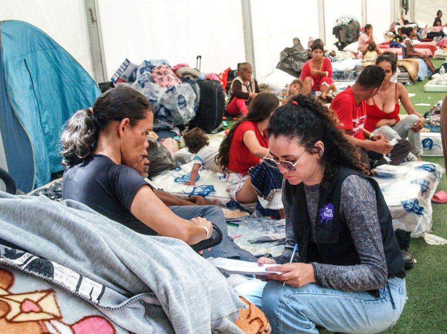 An aid worker talks with a woman in a temporary shelter. Many other earthquake survivors are visible in the background, sitting on mattresses on the floor.