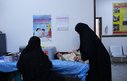 A healthcare worker checks a young child in a hospital room, watched by another woman who is likely the child's mother