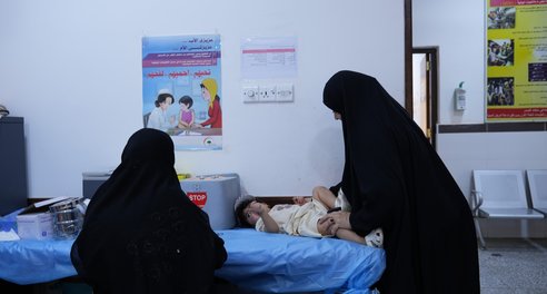 A healthcare worker checks a young child in a hospital room, watched by another woman who is likely the child's mother