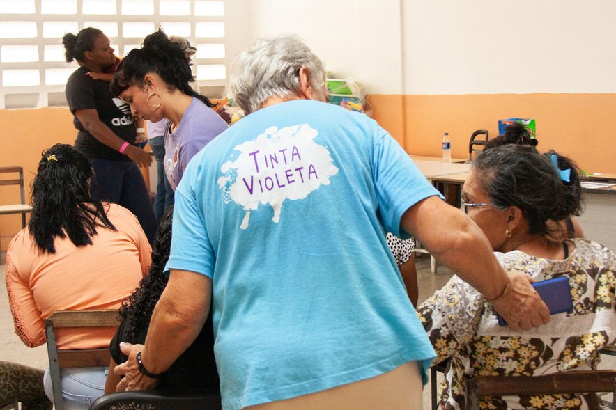 A woman wearing a t-shirt that says 'Tinta Violeta' talks with other women gathered in a meeting room