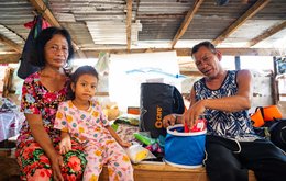 A woman, man and young girl sit in a makeshift shelter, in the process of pulling out items from an aid package.