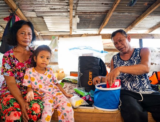 A woman, man and young girl sit in a makeshift shelter, in the process of pulling out items from an aid package.