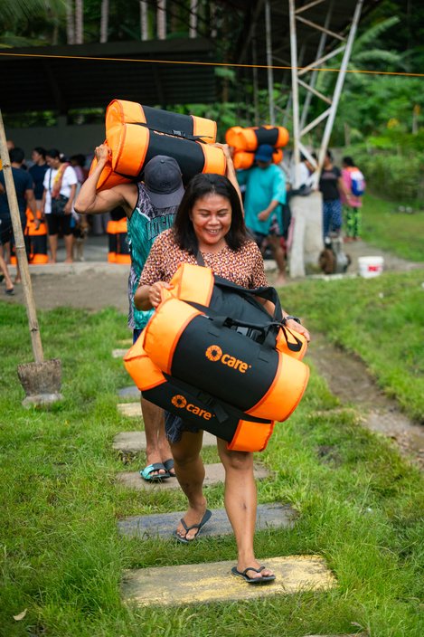 A woman walks along holding a black and orange package with the CARE logo on it. Several other people are visible in the background carrying similar packages on their backs.