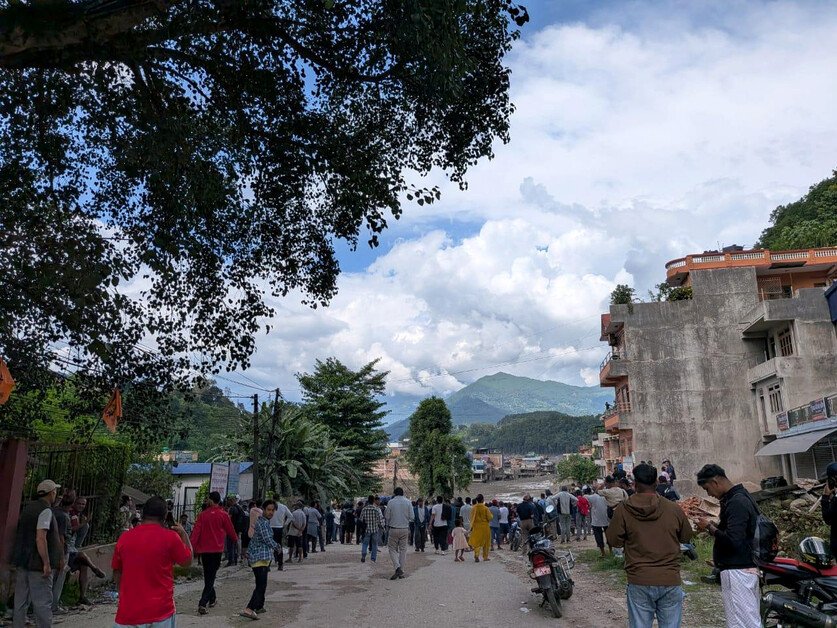 A crowded street scene in a mountainous region. A large group of people are walking along the road, flanked by buildings and motorcycles.