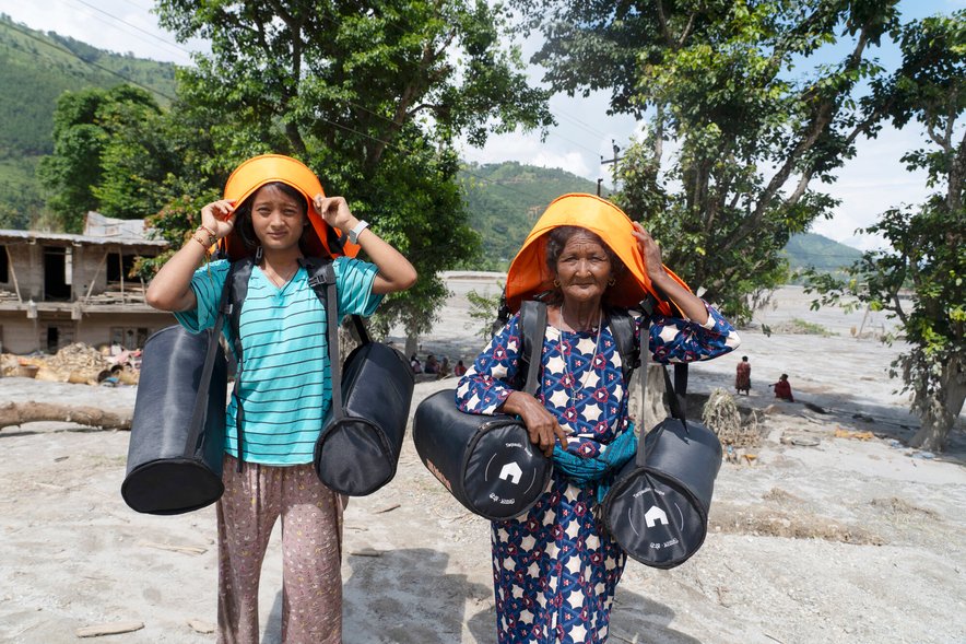 Two women walk with a barren landscape visible in the background, they are carrying emergency supply kits which have an orange band going over their heads to help with carrying the kits.
