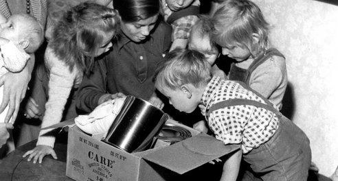A black and white image of six young children leaning over a cardbox box full of food