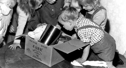 A black and white image of six young children leaning over a cardbox box full of food