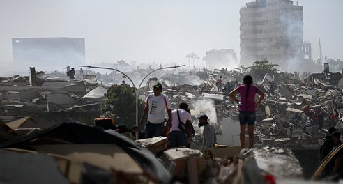 A scene of devastation following an earthquake - piles of rubble and collapsed buildings. Several people are visible searching through the rubble
