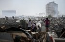 A scene of devastation following an earthquake - piles of rubble and collapsed buildings. Several people are visible searching through the rubble